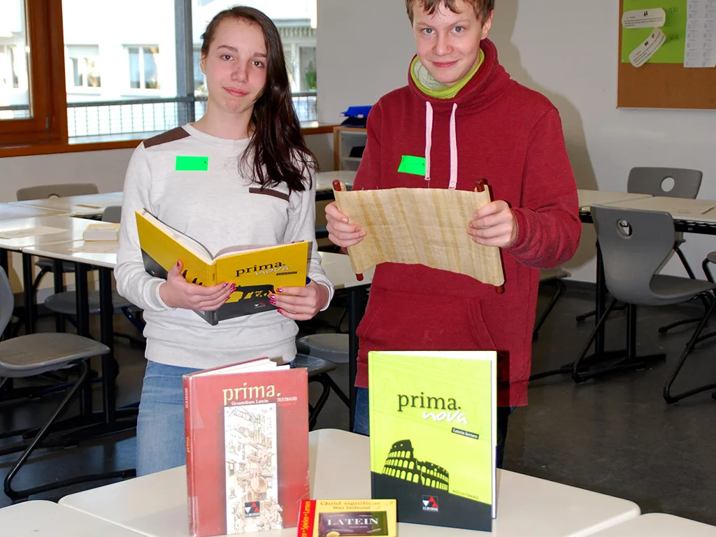 Zwei Jugendliche zeigen an einem Tisch Lehrbücher für Latein.
